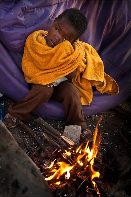 A child wrapped in a blanket, sitting beside a fire